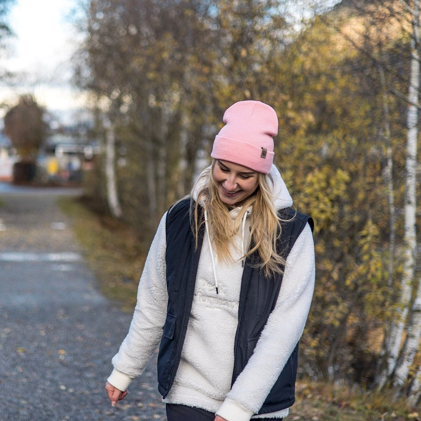 A young woman with long blonde hair smiles while looking downward. She is wearing a light pink Color Beanie by Goggleface, a grey hoodie, and a black vest made from recycled materials. She stands on a path lined with trees showcasing vibrant autumn foliage. The background is slightly blurred, suggesting a nearby residential area.