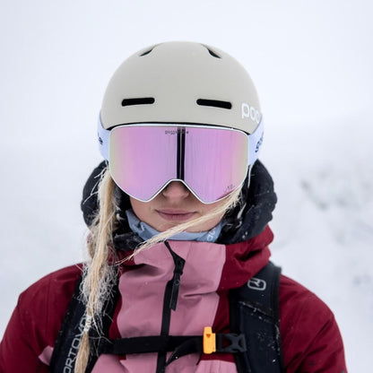 A person wearing the Goggleface Straight Line | White jacket and a helmet with pink-tinted snow goggles stands in a snowy environment. Strands of blonde hair are visible under the helmet, blending perfectly with the white, snowy background—a true Goggleface in winter wonderland.